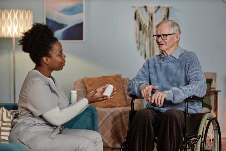A caregiver assists an older adult with hand hygiene in a well-lit common area of a residential care community.